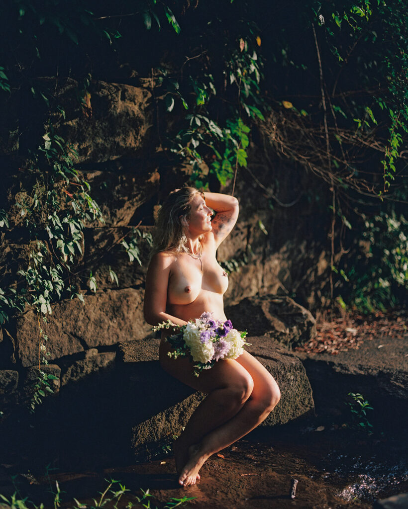 Nude woman wrapped in bouquet of flowers sitting on textured rocks, outdoor fine art film photography by K Hammock Photography North Carolina