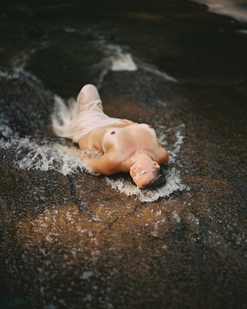 Woman wrapped in sheer fabric laying in running creek, outdoor fine art film photography by K Hammock Photography North Carolina