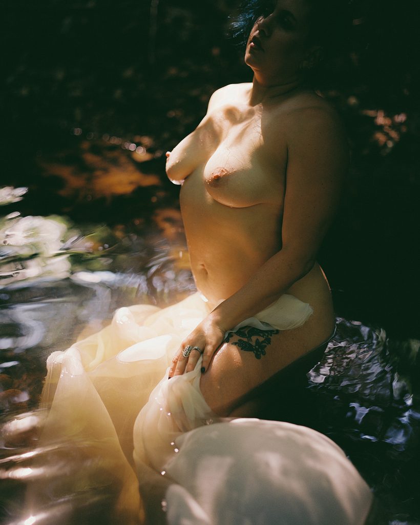 Woman sitting in a sunlit creek with sheer ivory fabric draped across her lap during an outdoor film boudoir session.