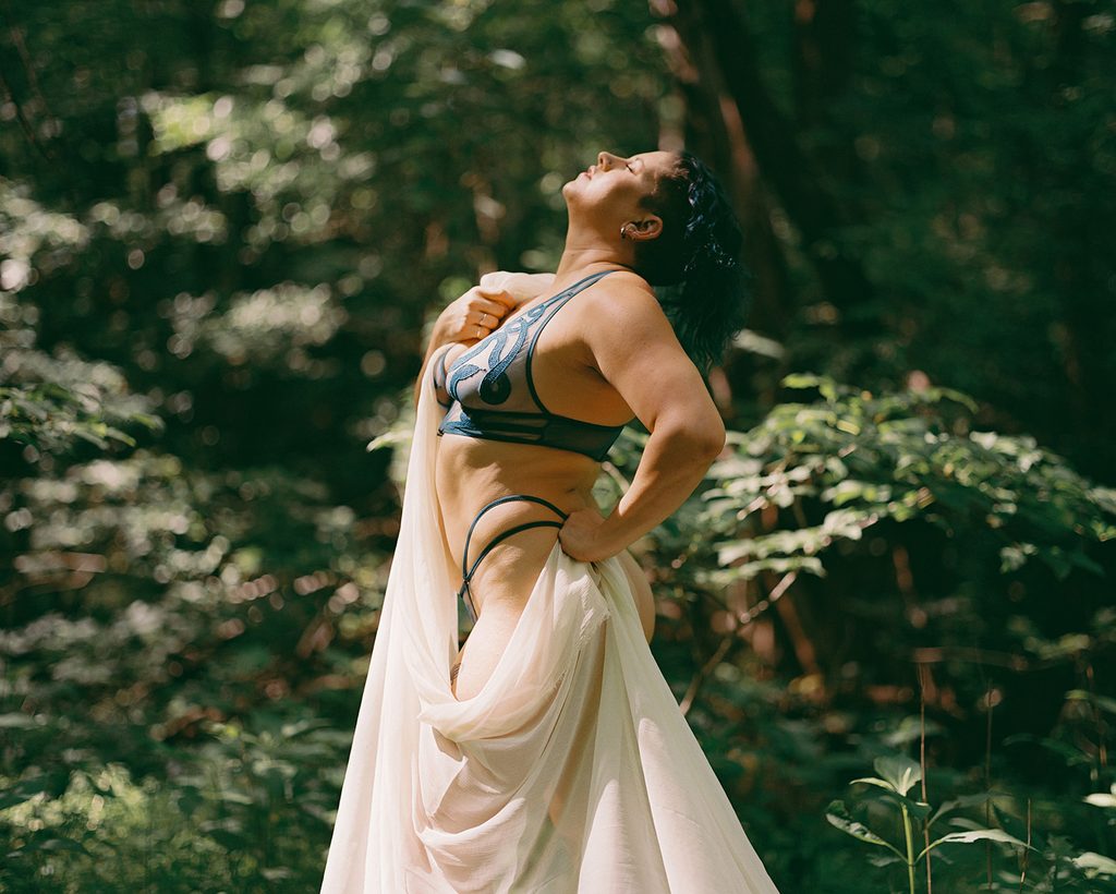 Woman wearing blue lingerie with flowing ivory fabric standing in a lush green forest with her face tilted toward the sunlight.