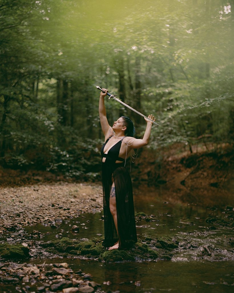 Woman standing barefoot in a shallow forest creek wearing black lingerie and holding a sword above her head.