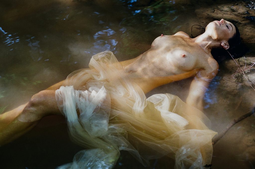 Woman reclining in shallow creek water with sheer ivory tulle flowing around her during a fine art film boudoir session.