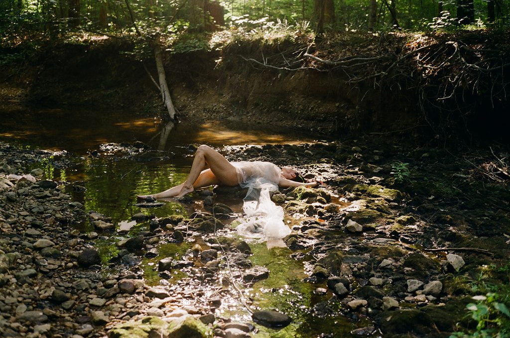 Wide film photograph of a woman reclining among rocks in a shallow forest creek with sheer white fabric draped around her.