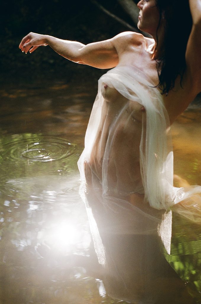 Woman standing in a sunlit creek with sheer ivory fabric draped around her body during an outdoor film boudoir session.