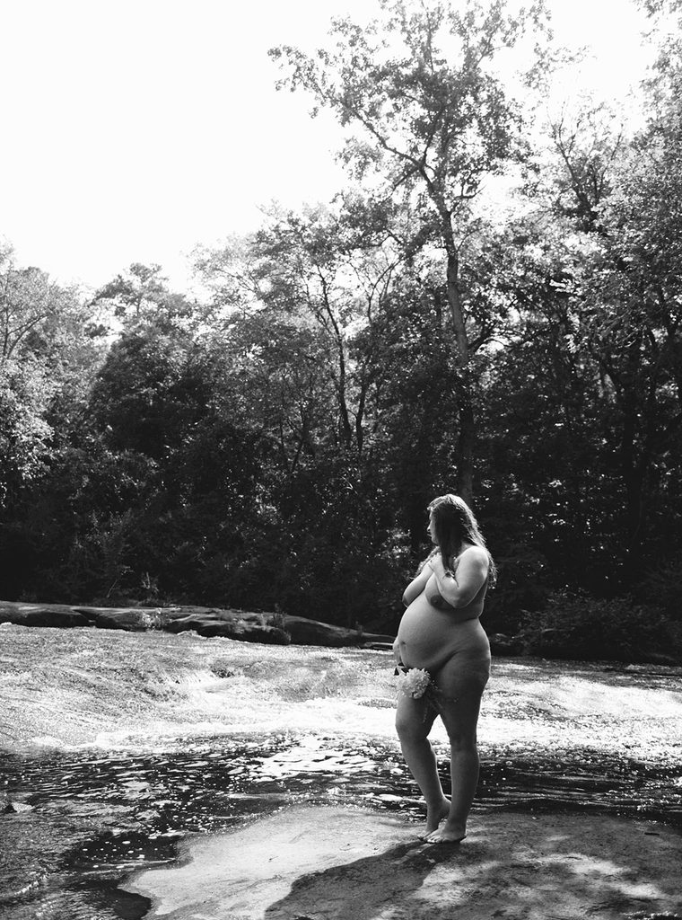 Black and white outdoor maternity boudoir portrait of a pregnant woman standing barefoot beside a flowing creek.