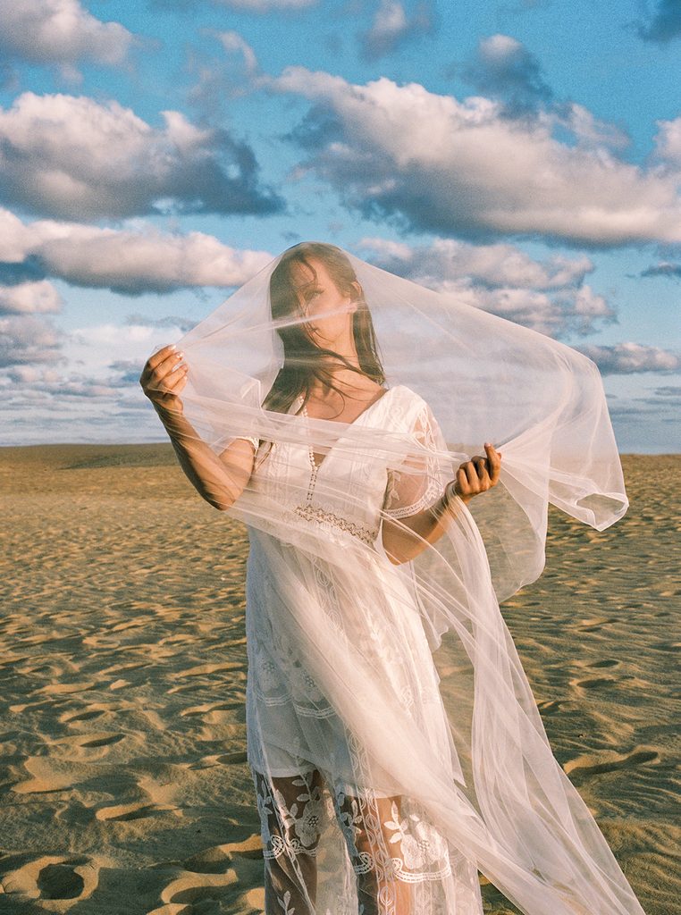 Woman holding a sheer white veil around her face while standing on sunlit sand dunes during an outdoor boudoir photography session.