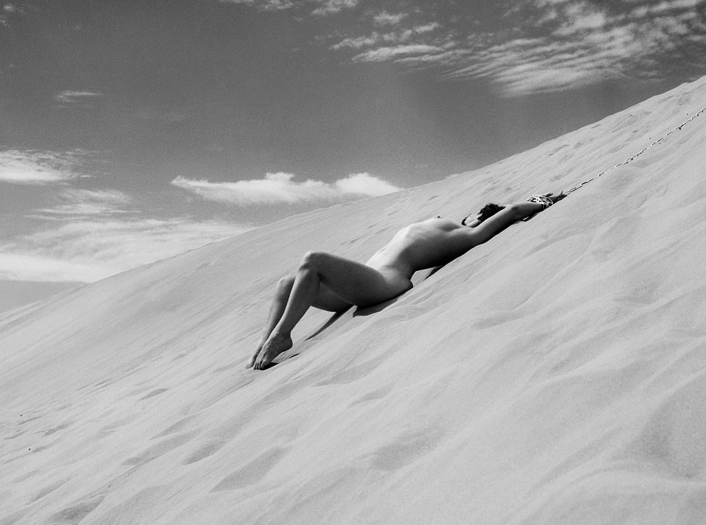 Black and white film boudoir photograph of a nude woman reclining on a sloping sand dune beneath an open sky.