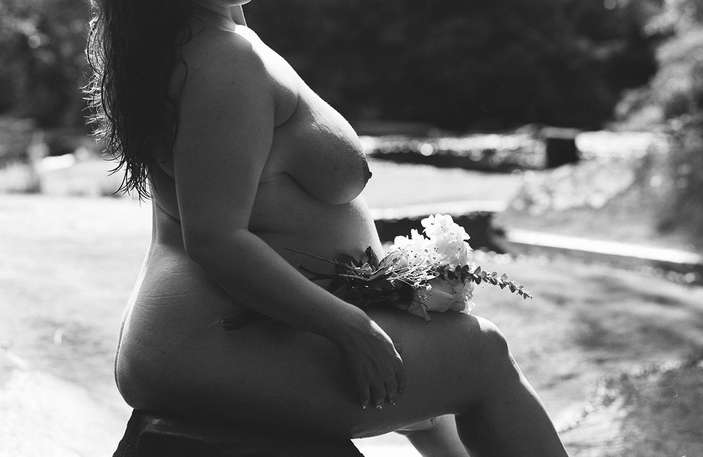 Black and white fine art boudoir portrait of a nude pregnant woman seated outdoors with a bouquet of white flowers resting on her lap.