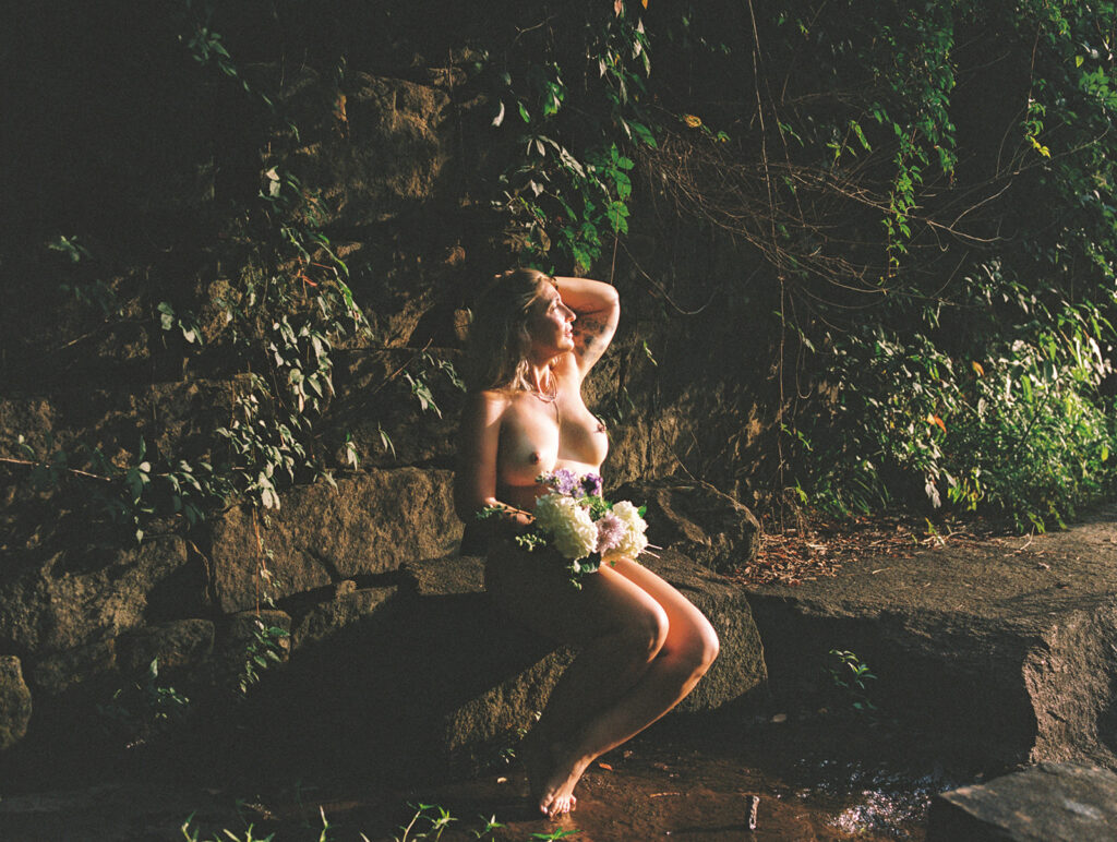 Woman seated on rocks beside a wooded creek, holding flowers during an outdoor boudoir session.