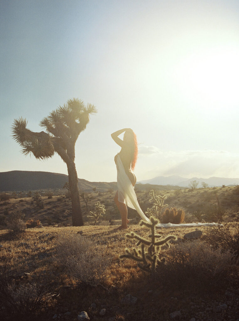 Black-and-white film portrait of a woman in a sheer white dress leaning against a desert tree.