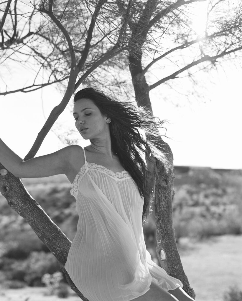 Woman draped in white fabric standing beside a Joshua tree in warm desert sunlight.
