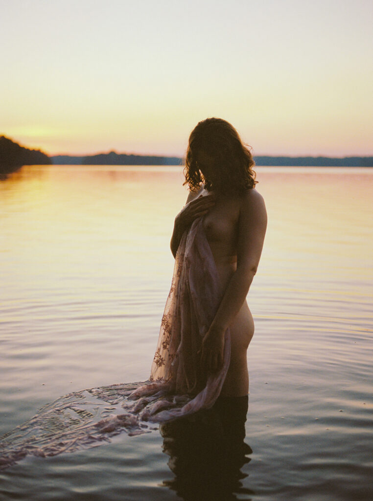 Woman standing waist-deep in a lake at sunset, draped in lace during an outdoor boudoir session.