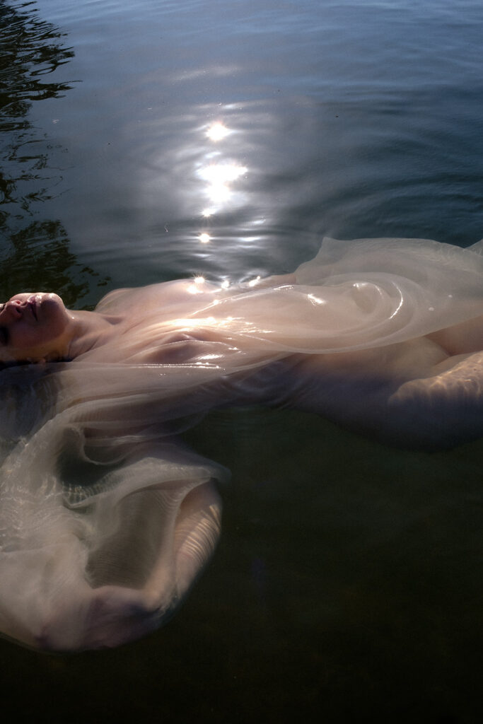 Woman floating in calm water as sunlight reflects across sheer white fabric during a Goddess Session.