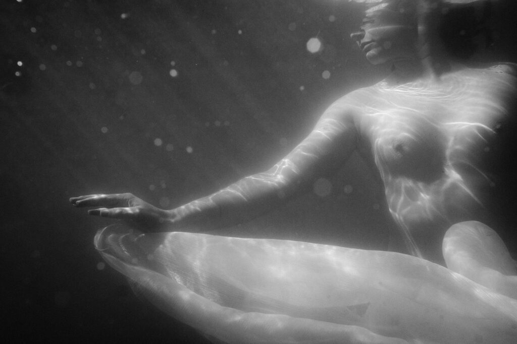 Black-and-white underwater boudoir portrait with light moving across a woman’s body and flowing fabric.