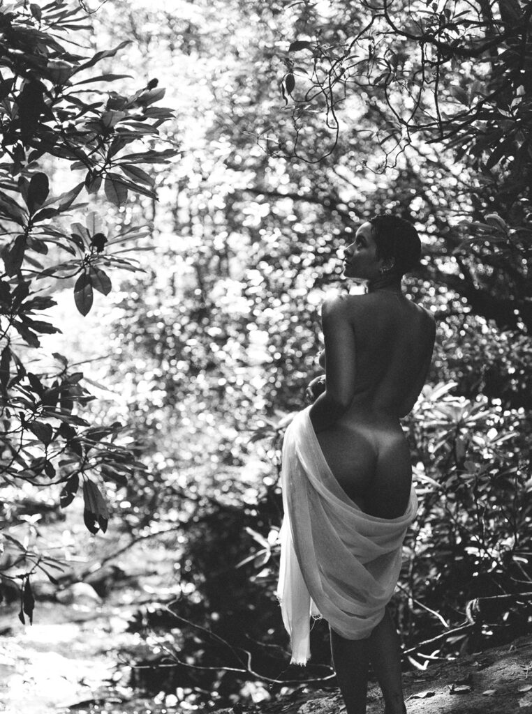 Black-and-white film boudoir portrait of a woman draped in white fabric beside a wooded creek.