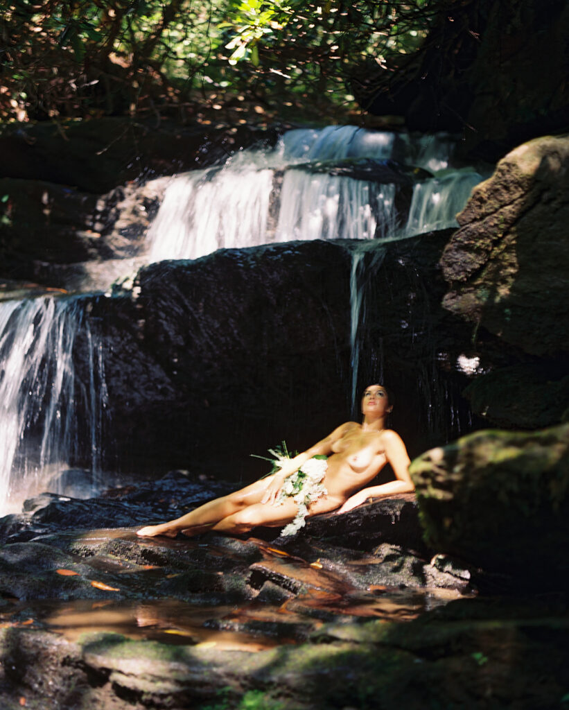 Woman reclining with flowers beneath a waterfall during a nature-inspired Goddess Session.
