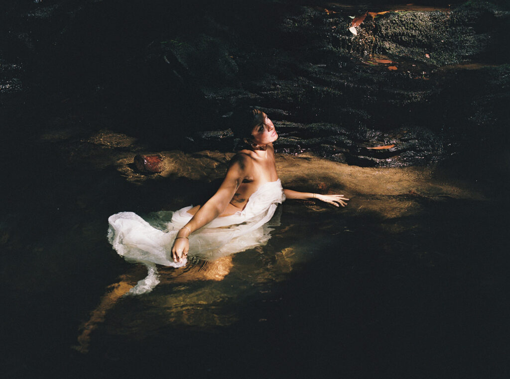 Woman wrapped in flowing white fabric sitting in shallow creek water during an outdoor boudoir session.